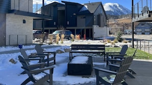 A modern outdoor seating area with Adirondack chairs and a fire pit, surrounded by a building with a view of a snow-capped mountain.