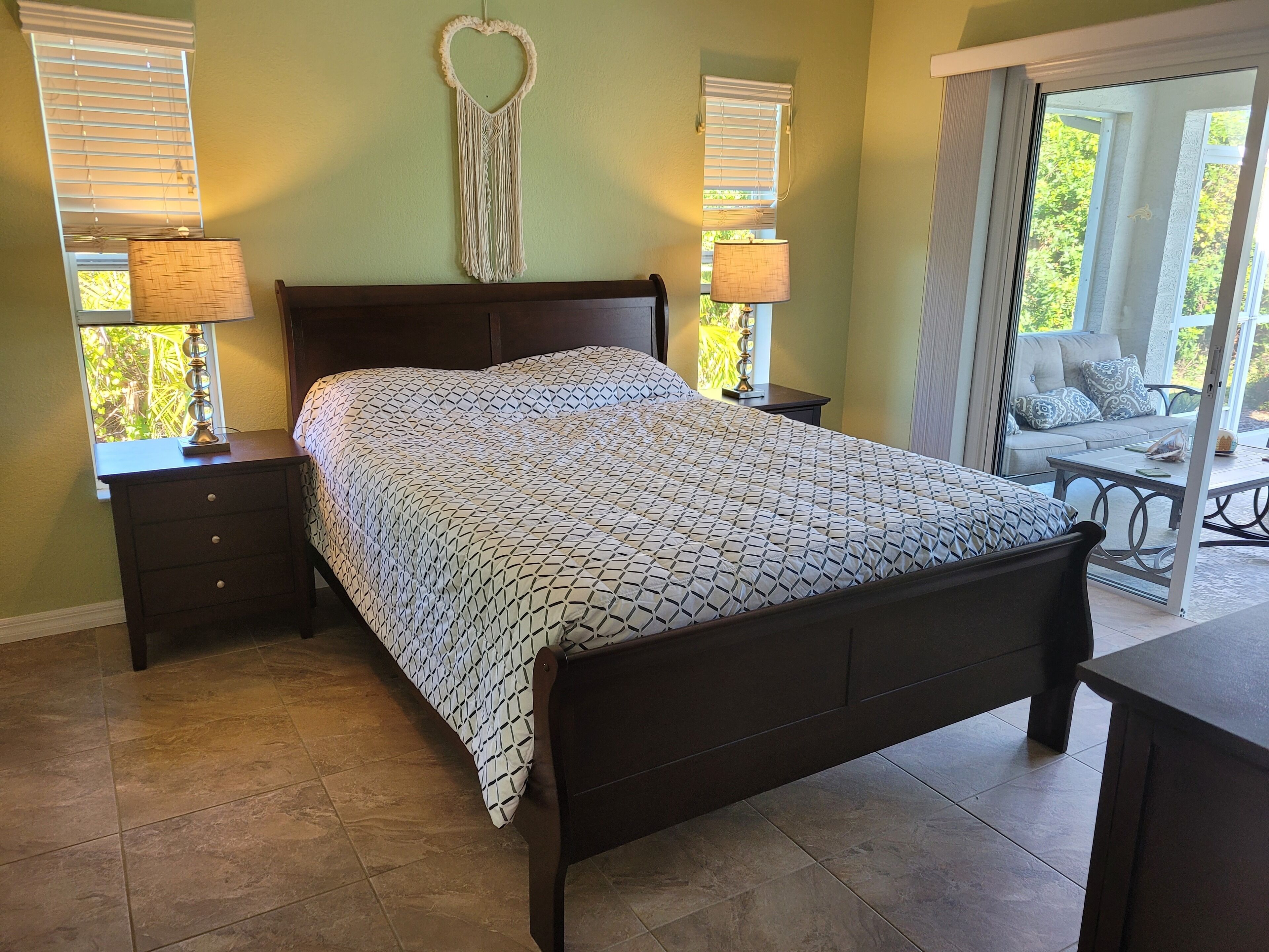 A bedroom with a bed, two bedside tables with lamps, a window with blinds, and a view of a balcony.