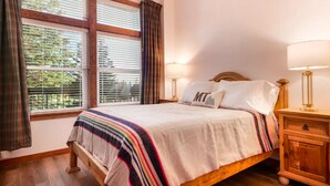 Un dormitorio con una cama, dos mesitas de noche, una ventana con persianas y cortinas.