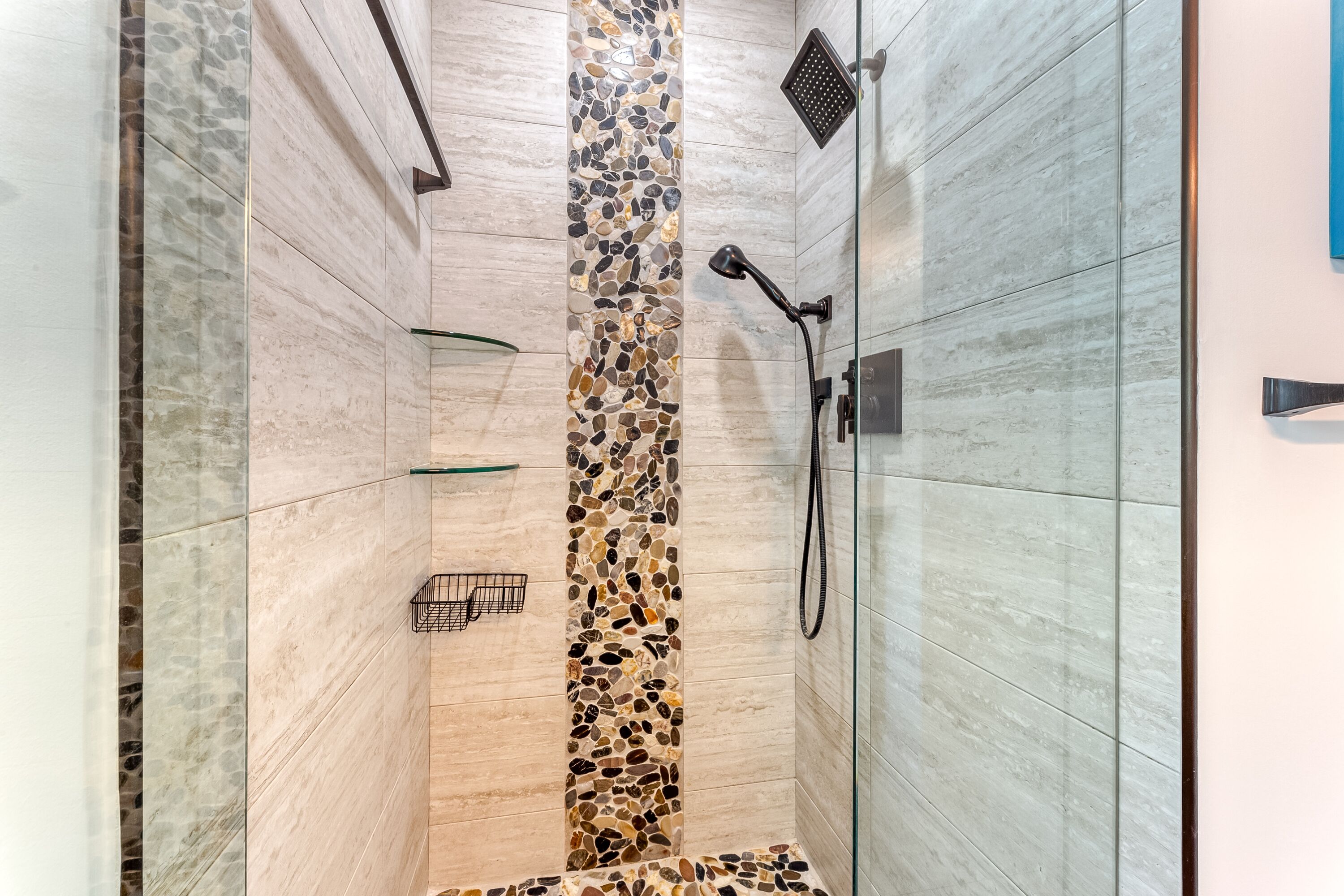 Une douche fermée par des parois de verre, avec un mur décoré d’une mosaïque de galets, une étagère en verre et une douchette à main.