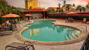 Una piscina con sillas de descanso y sombrillas, rodeada de palmeras y un edificio al fondo.