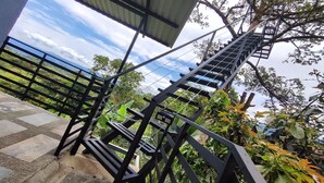 Una escalera con barandilla de metal que sube hacia un espacio cubierto, rodeado de abundante vegetación y vistas a colinas distantes.