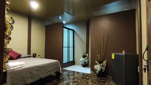 Habitación de hotel con cama, mesita de noche, lámpara, un jarrón con ramas decorativas y un refrigerador.