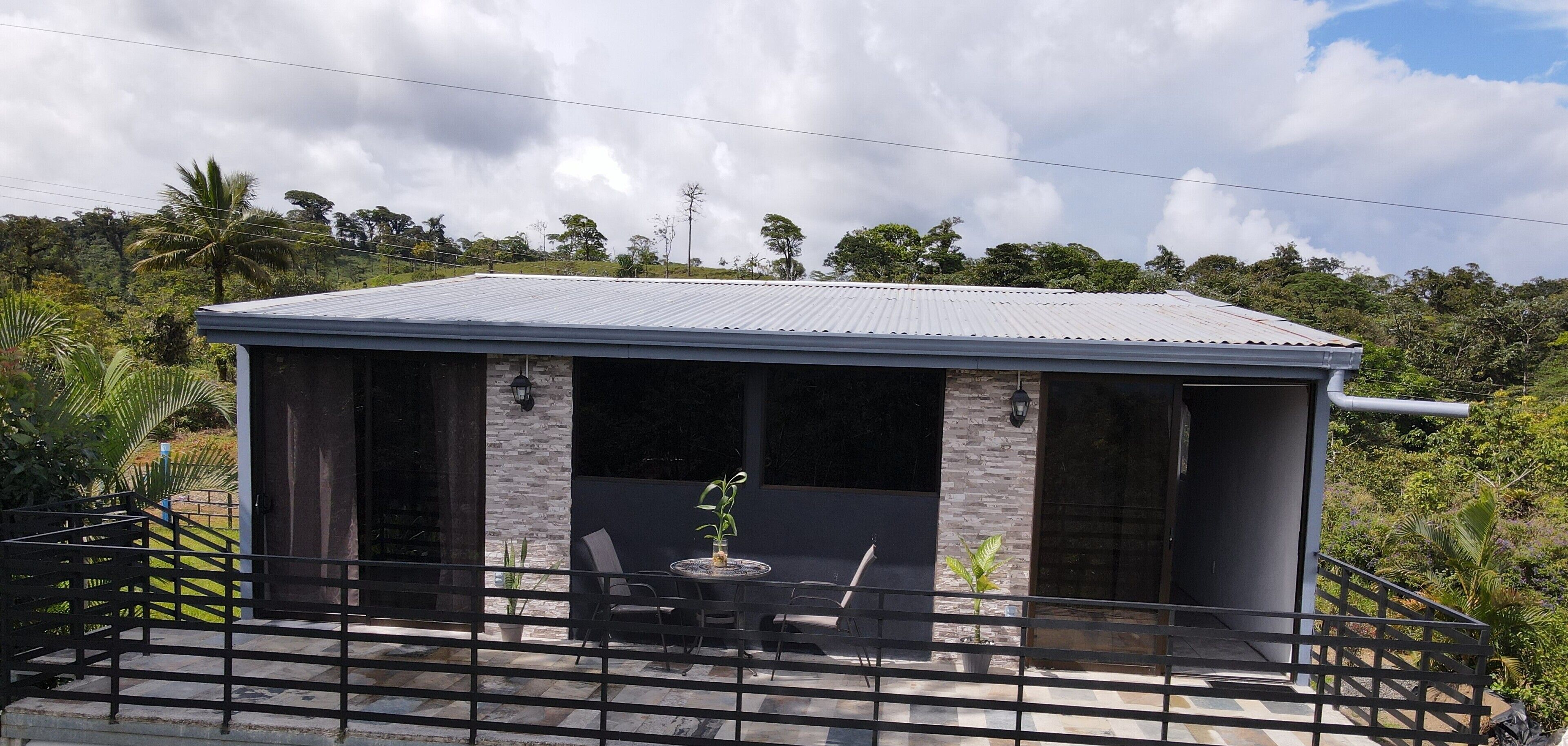 A small, single-story building with a metal roof and stone walls, featuring a patio with a table and chairs, surrounded by lush greenery.