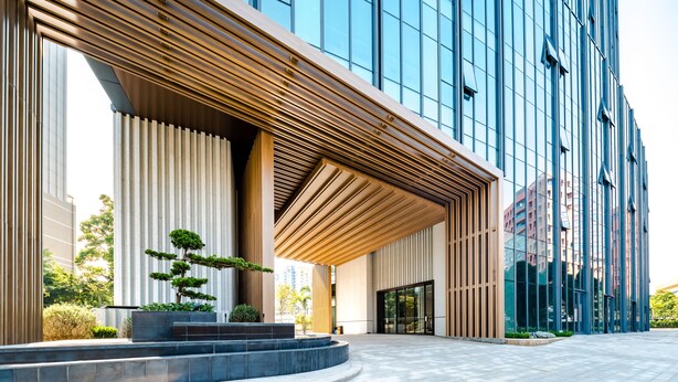 Un edificio moderno con un amplio frente de vidrio, un alero de madera y un área ajardinada con un pequeño árbol.