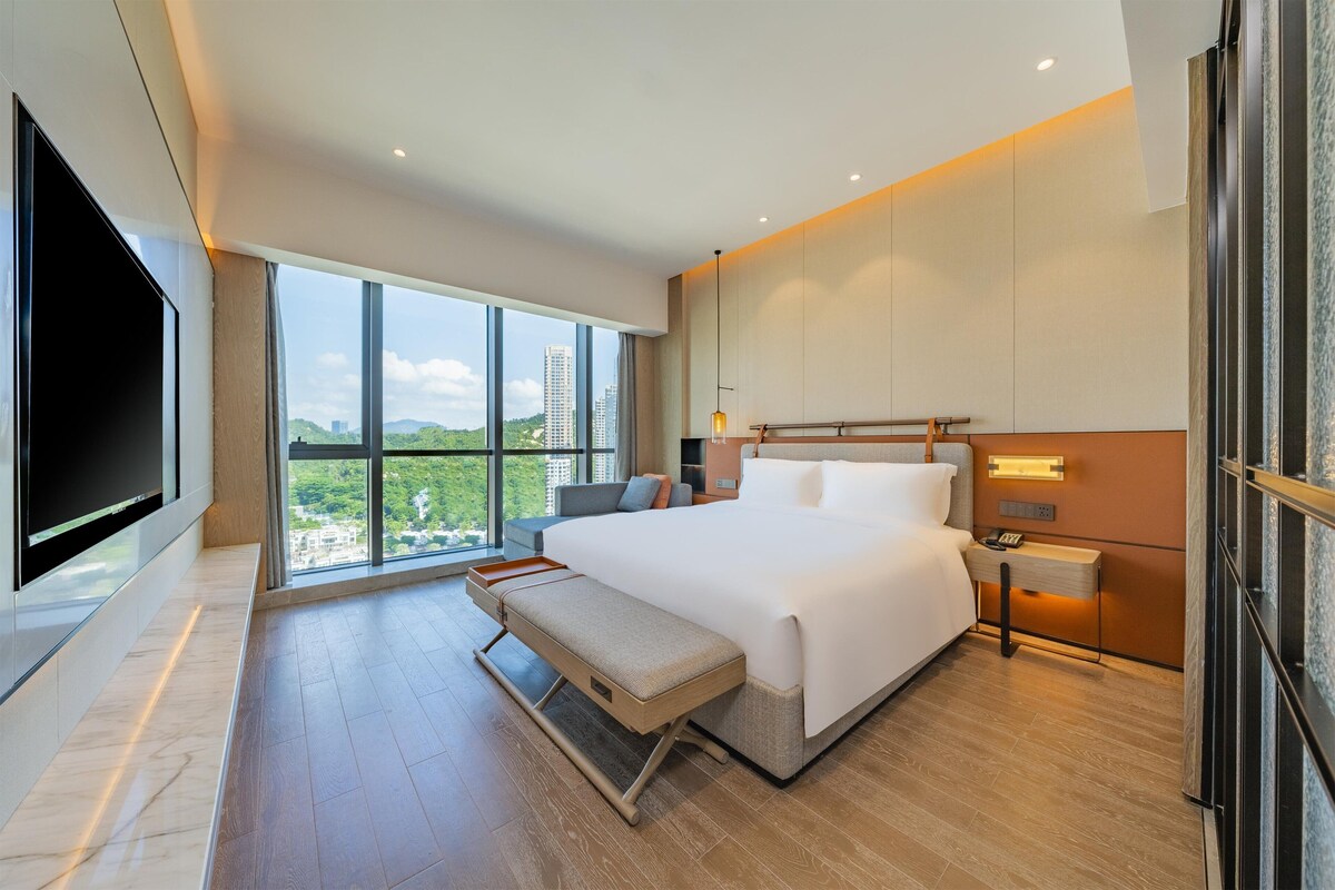 A modern hotel room with a large bed, a bench, a TV, and a view of the city.