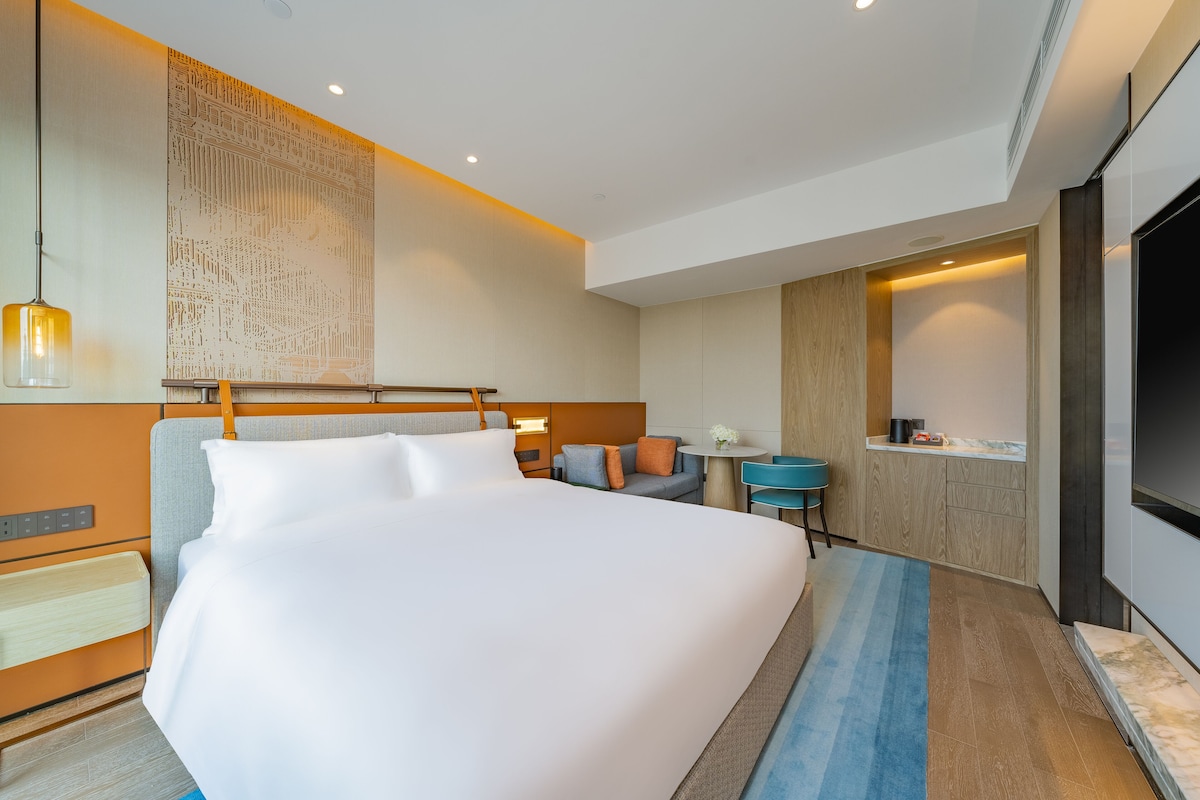 A modern hotel room with a large bed, a desk, a TV, and a wall-mounted artwork.