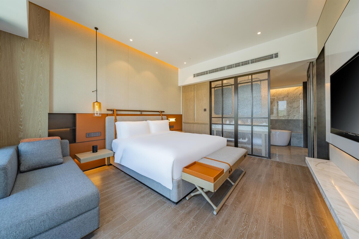 Een moderne hotelkamer met een groot bed, een bank, een televisie en een badkamer zichtbaar door een glazen deur.