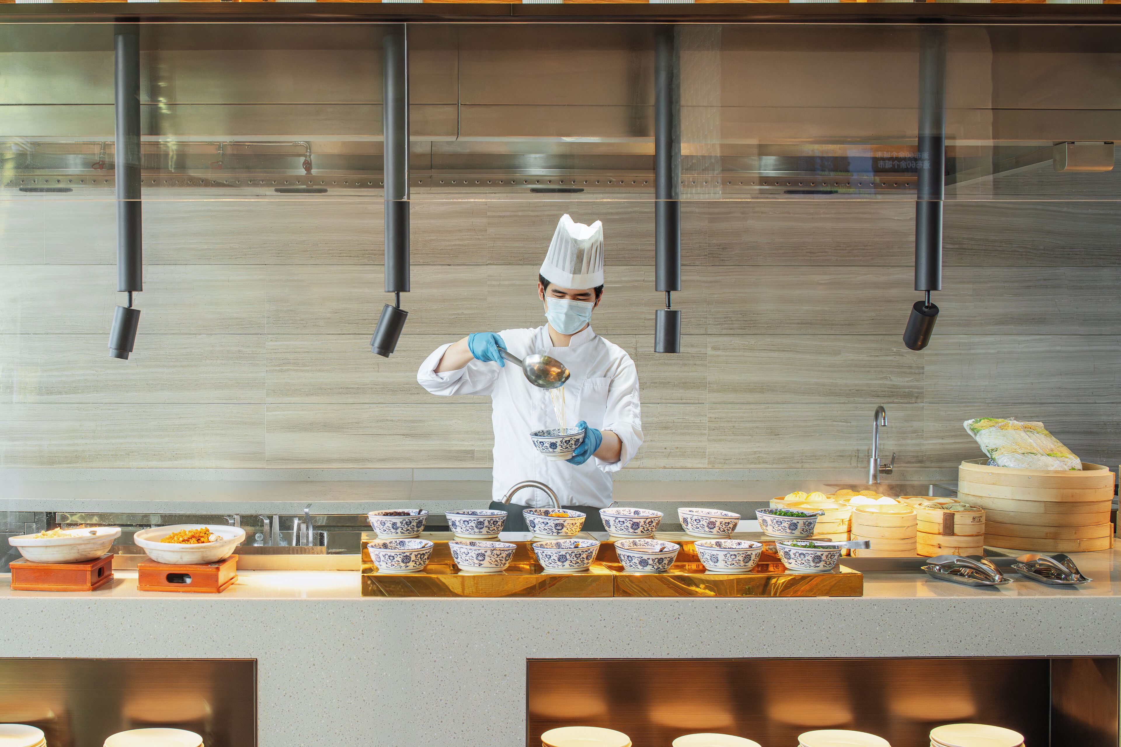 Ein Koch in weißer Uniform und Kochmütze bereitet Essen in einer Küche mit Edelstahlgeräten und einer gefliesten Spritzwand zu.