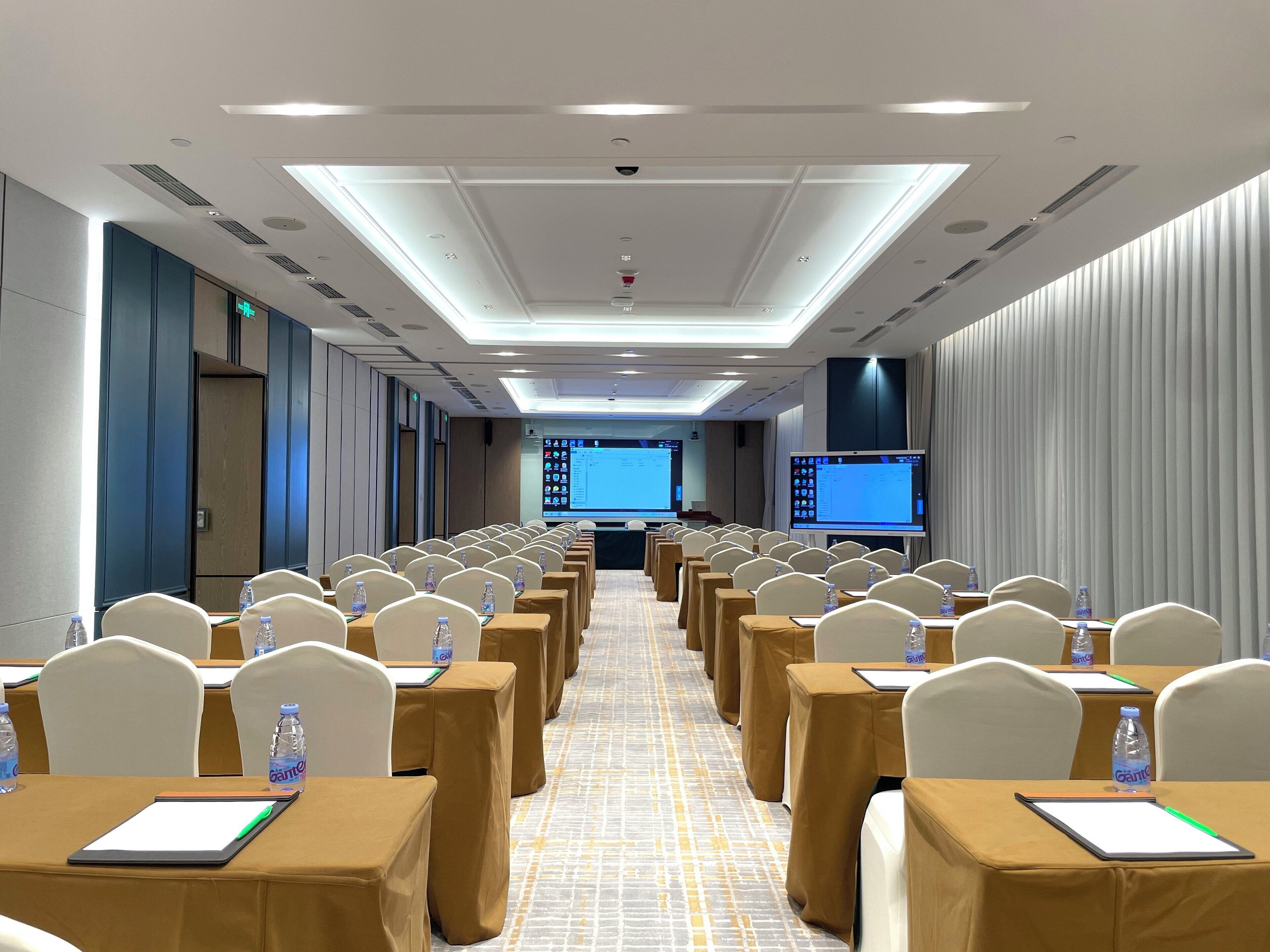 Un salón de conferencias con filas de mesas, sillas y una pantalla grande al frente.