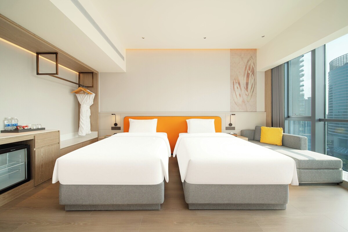 A modern hotel room with two single beds, a sofa, a desk with a refrigerator, and a large window offering a city view.