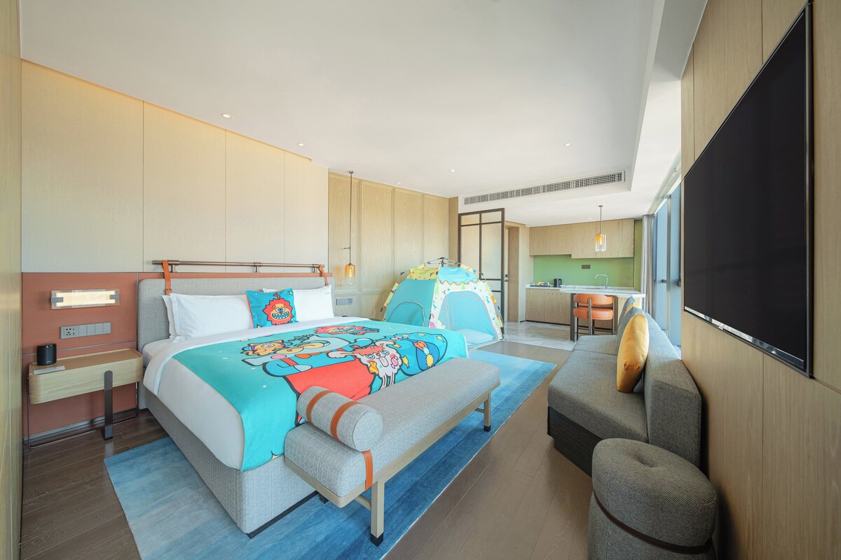 A modern hotel room with a large bed, a bench, a small sofa, a desk, and a large TV screen.