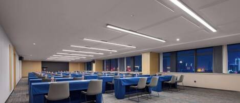 A conference room with multiple tables and chairs, large windows, and a carpeted floor.