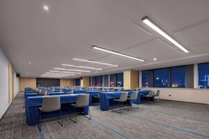 Un salón de conferencias con varias mesas y sillas, ventanas amplias y piso alfombrado.