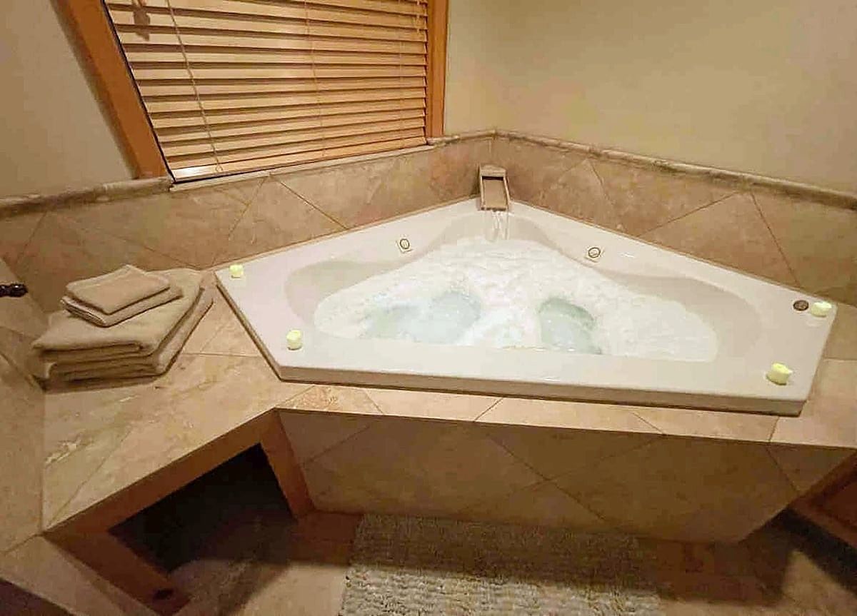 A corner bathtub with a built-in seat, a towel stack, and a window with blinds.