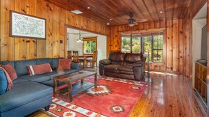 A spacious living room with wooden walls, a large window, a ceiling fan, and a dining area with wooden furniture. - 🛶 Cozy Creek Cabin near Yosemite & Bass Lake (Ahwahnee)