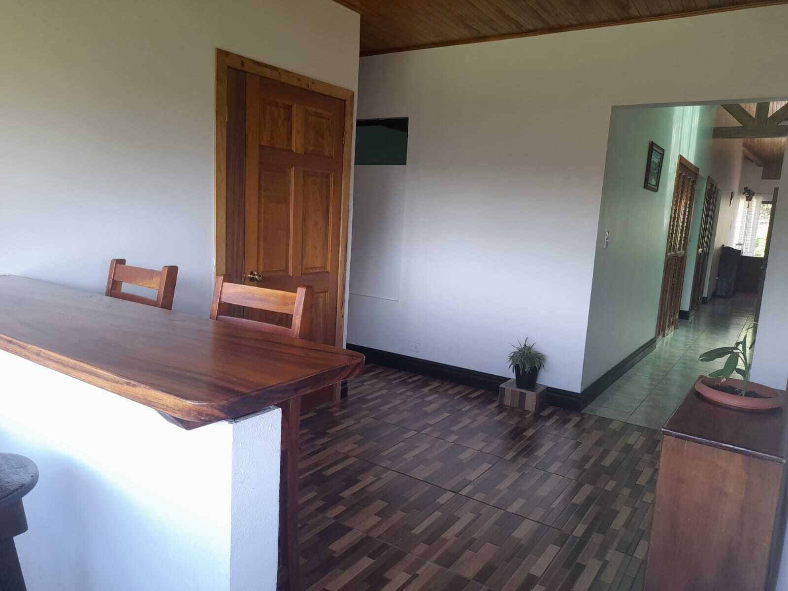 A dining area with a wooden table and chairs, a small potted plant, and a hallway with a wooden door and a light switch on the wall.