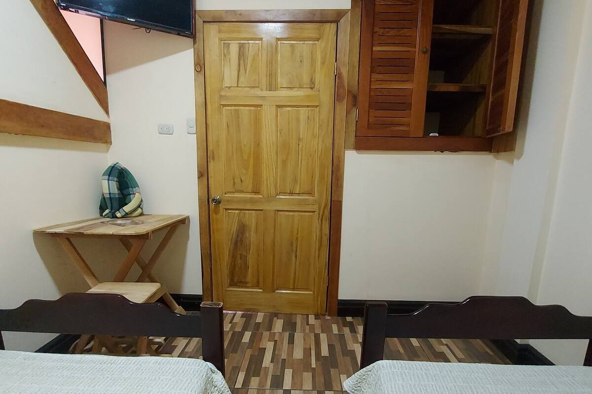 A room with two beds, a wooden door, a TV mounted on the wall, and a small table with a chair.