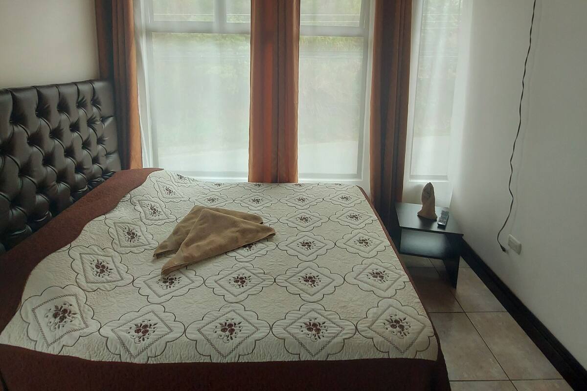 A bedroom with a large bed, wooden ceiling, and two windows with curtains.