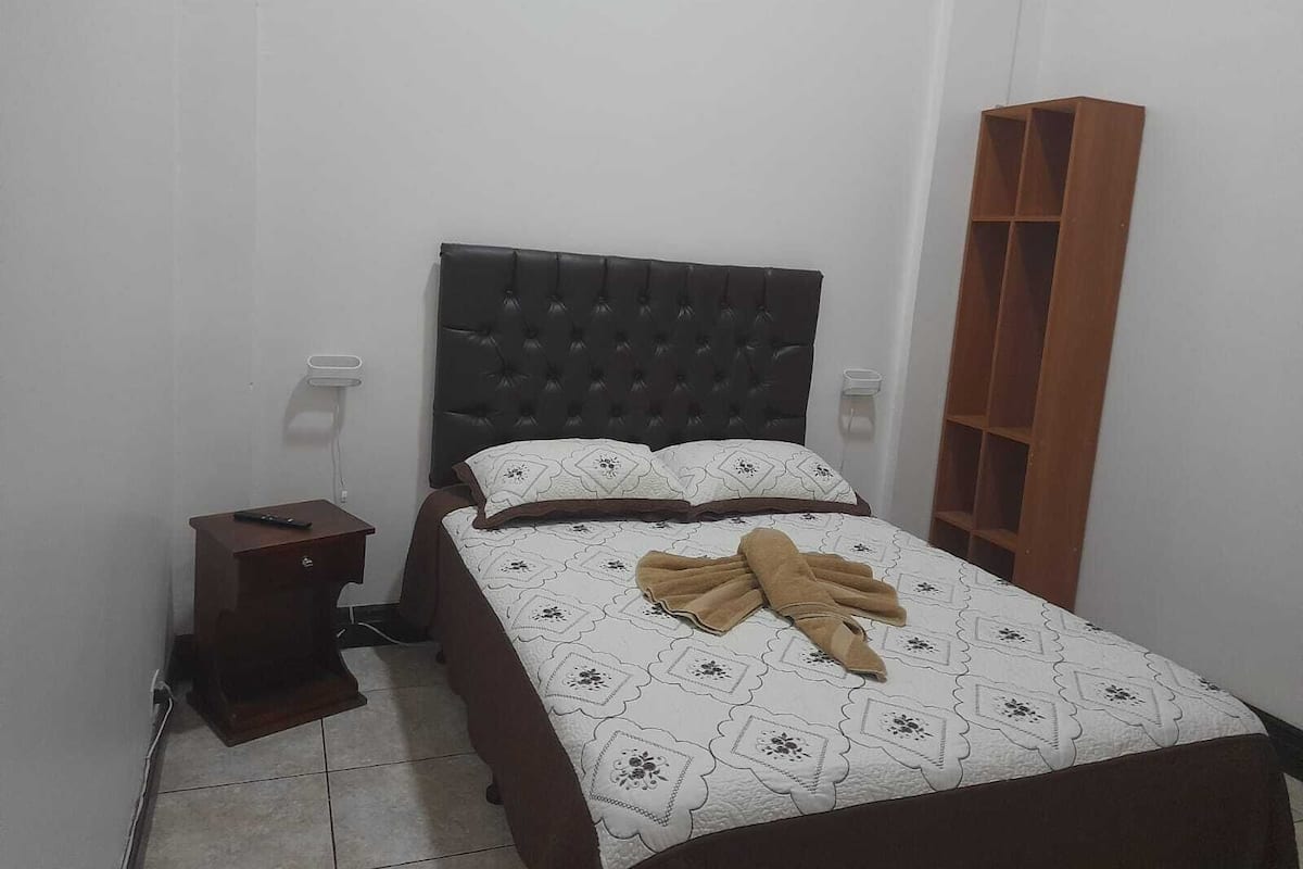 A bedroom with a bed, a nightstand, a wooden shelf, and a wall socket.