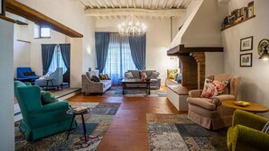 A spacious living room with a high ceiling, exposed wooden beams, a chandelier, and a fireplace. There are multiple seating areas with sofas and armchairs, and a bookshelf filled with books.