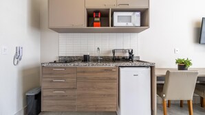 Cocina compacta con microondas, un refrigerador pequeño y un lavamanos. Se ve un cubo de basura y una silla.