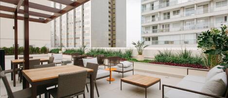 An outdoor patio with tables and chairs, surrounded by greenery and overlooking high-rise buildings.