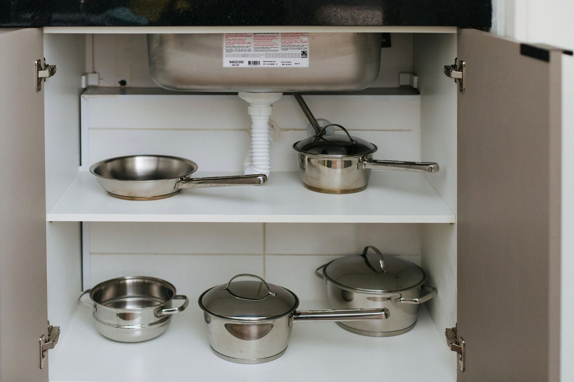 A kitchen cabinet with stainless steel cookware including a frying pan, two pots, and two pans.