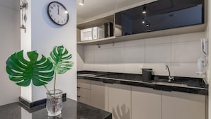 A modern kitchen with white cabinets, a black countertop, a clock, a microwave, and a potted monstera plant. - Av0111 Studio Avanhandava (São Paulo)