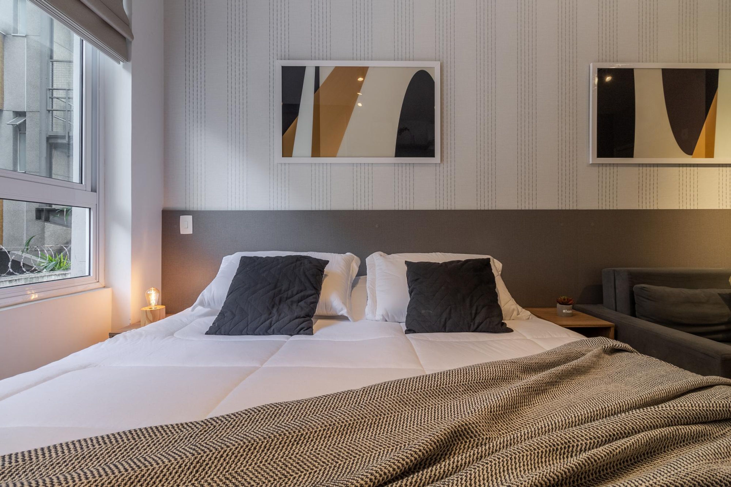 A neatly made bed with white and black bedding, a patterned throw blanket, and two abstract paintings on the wall above.