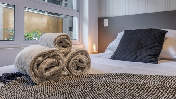 A neatly made bed with rolled towels and a patterned throw, a bedside lamp, and a window with a view of the outside. - Av0111 Studio Avanhandava (São Paulo)