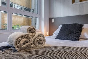 A neatly made bed with rolled towels and a patterned throw, a bedside lamp, and a window with a view of the outside. - Av0111 Studio Avanhandava (São Paulo)