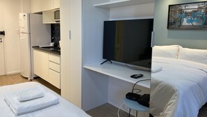 Une chambre d’hôtel moderne dotée d’une kitchenette, d’un téléviseur à écran plat, d’un lit avec du linge de maison blanc et d’un tableau représentant des bicyclettes accroché au mur.