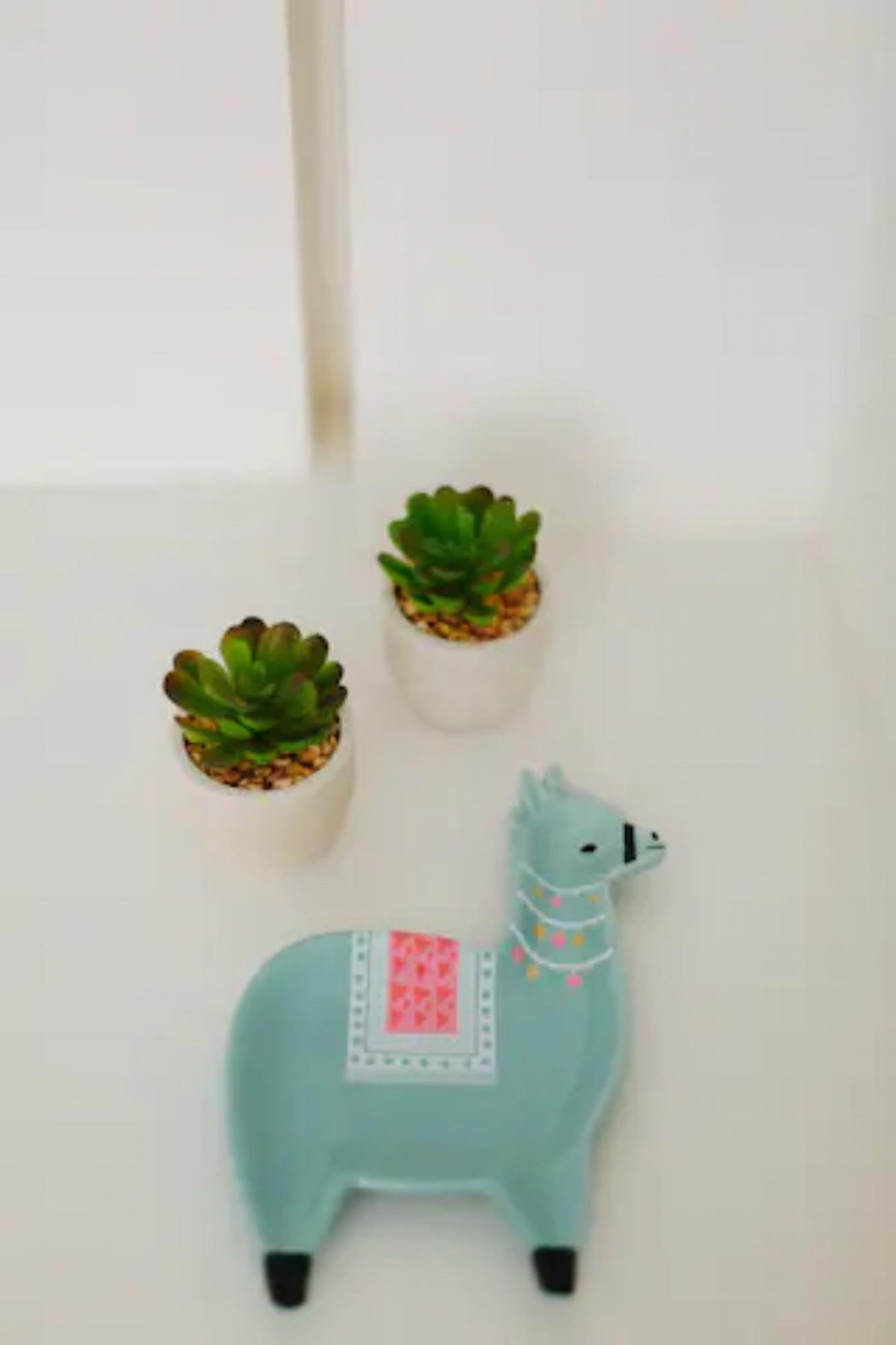 A small blue llama figurine with a pink and white patch on its back, positioned between two small potted plants on a white surface.