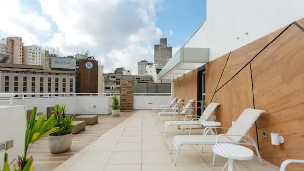 A rooftop terrace with white lounge chairs, potted plants, and a city view. - Av0408 Studio Avanhandava (São Paulo)