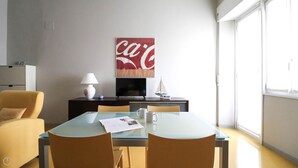 A modern hotel room with a glass dining table, two chairs, a sofa, a TV, and a large window.