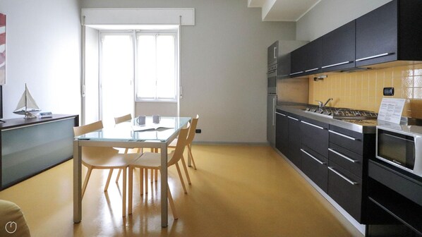A modern kitchen with dark cabinets, a gas stove, and a microwave. There is a dining table with chairs and a window providing natural light. - Dogali 17 - Santa Margherita Ligure (Santa Margherita Ligure)