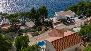 An aerial view of a coastal property with a swimming pool, a parking area, and a building with a red-tiled roof.