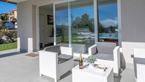 A modern outdoor patio with white wicker furniture, a glass door leading to a pool area, and a clear view of the surrounding landscape. - Villa Il Gelsomino Bellavista (Besozzo)