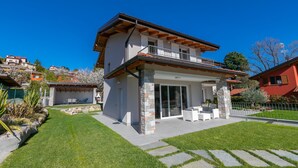 A two-story house with a balcony, a patio with outdoor furniture, and a well-maintained lawn. - Villa Il Gelsomino Bellavista (Besozzo)