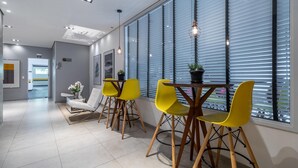 Un interior moderno con una zona de estar que incluye sillas amarillas y una mesa, ventanas amplias con persianas, y una puerta visible con la etiqueta 'SAIDA'.