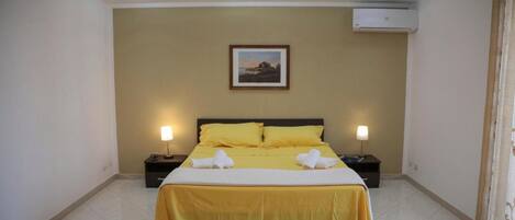 A hotel room with a bed, two bedside tables, a wall-mounted air conditioner, and a framed picture above the bed.