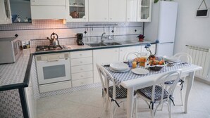 A kitchen with white cabinets, a gas stove, a sink, and a dining area with a table set for two. - Seascape Terrace Spacious Apartment With Terrace (Cefalù)