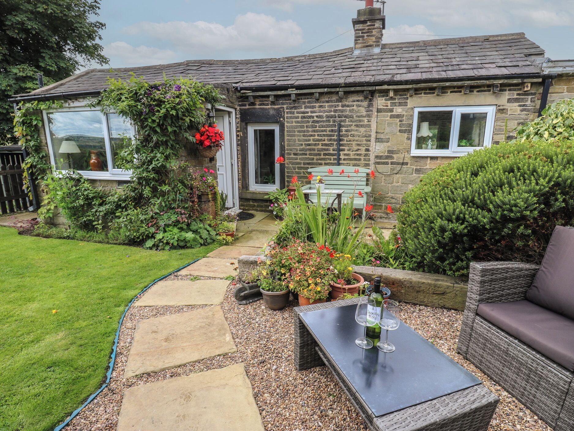 A stone house with a garden, a paved pathway, a wicker sofa, and a table with wine glasses.