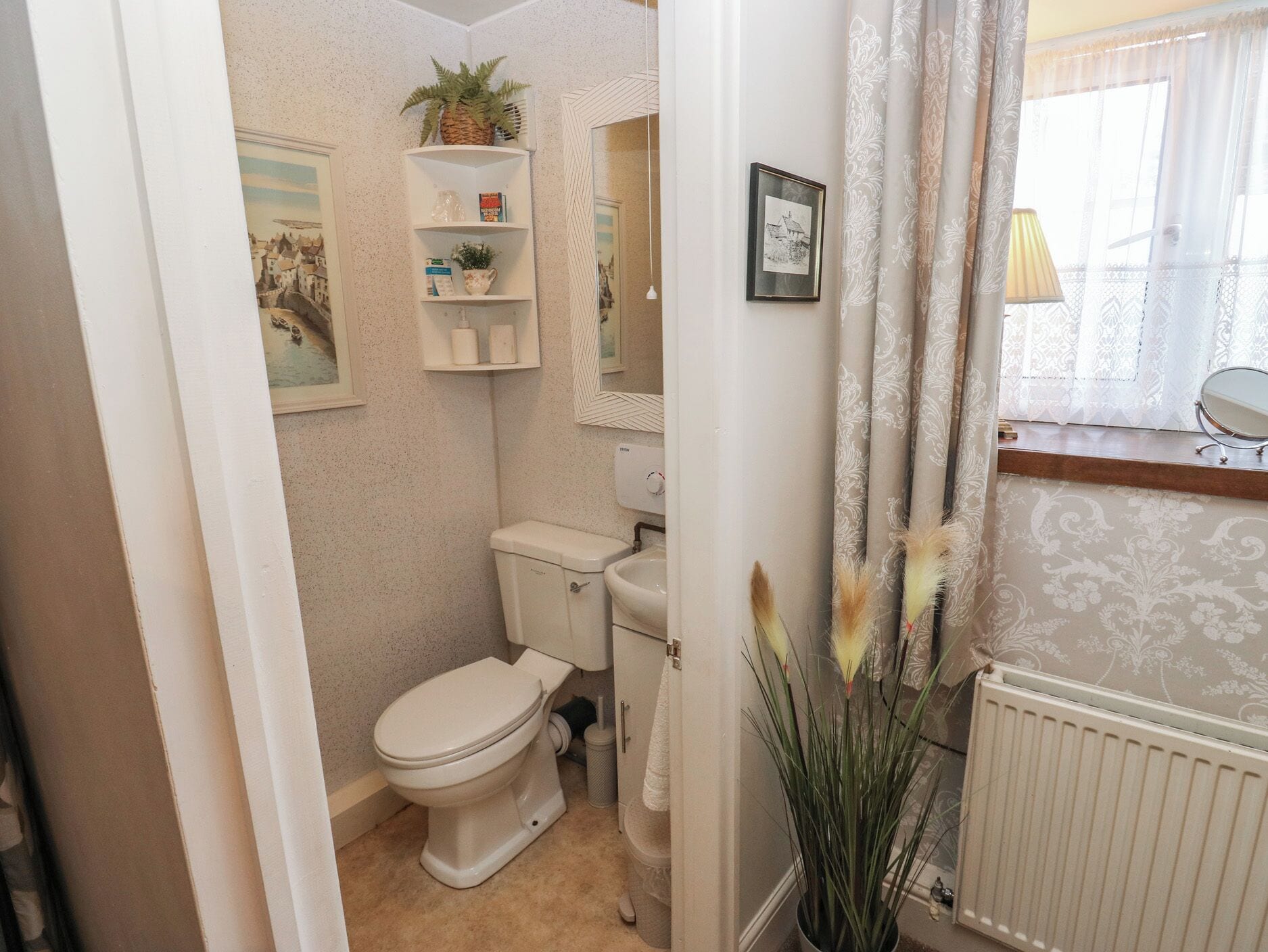 Baño pequeño y funcional con inodoro, lavabo y espejo. Objetos decorativos adornan la pared y hay una ventana con cortinas.