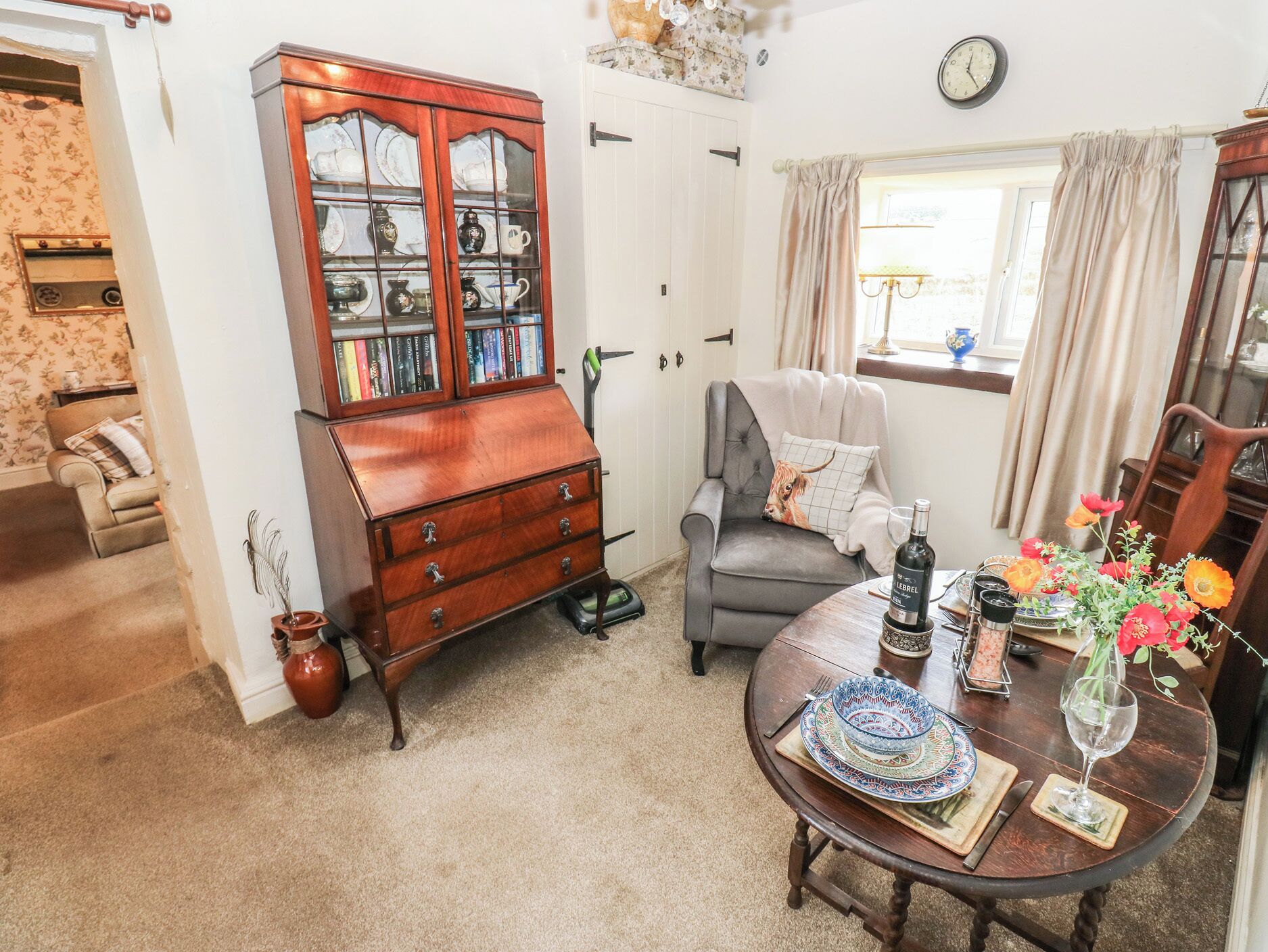 A cozy living room with a wooden cabinet, a grey armchair, a dining table with a vase of flowers, and a clock on the wall.