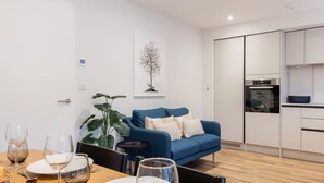 A modern living room with a blue sofa, dining table set with wine glasses and plates, a large plant, and a kitchen with white cabinets and appliances. - Driftwood (Newquay)
