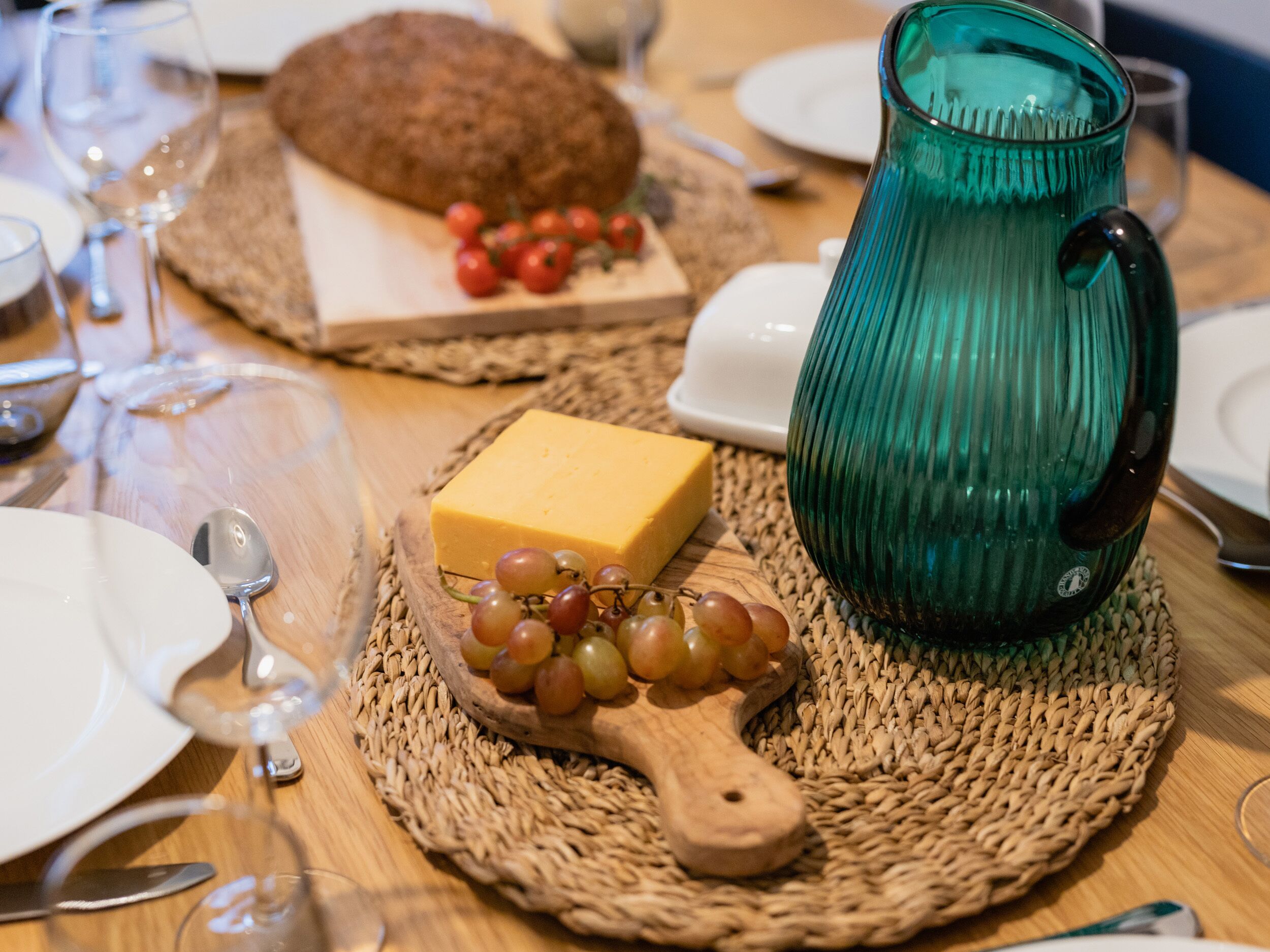 Une table avec un set de table en fibres tressées, une cuillère en bois accompagnée de raisins, un morceau de fromage, une miche de pain et un pichet en verre vert.
