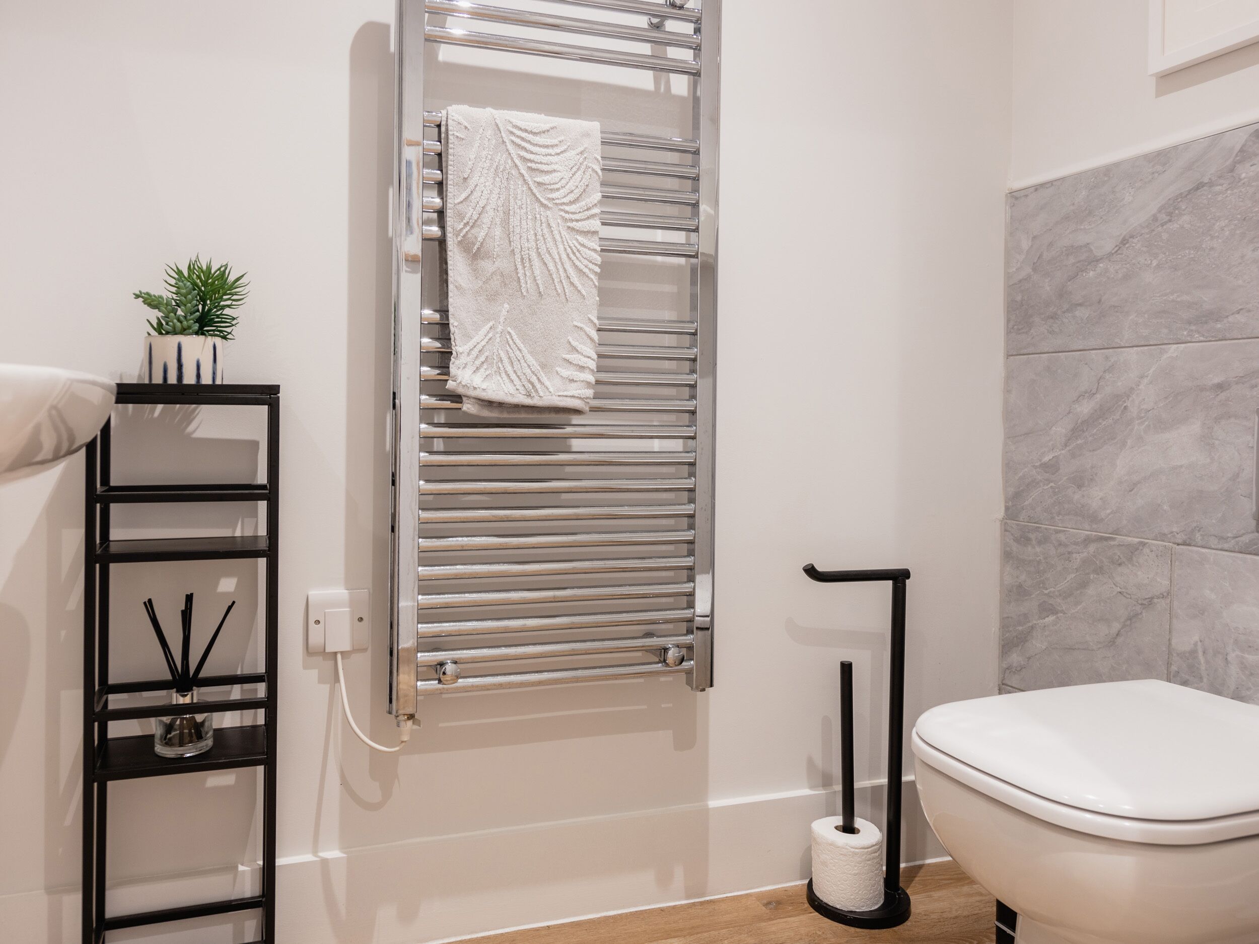 Et moderne badeværelse med et hvidt toilet, et håndklædestativ med et hvidt håndklæde, en sort hylde med en plante, og en væg i marmor.