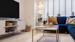 A modern living room with a flat-screen TV, a blue sofa, a coffee table, and a large window with blinds. - Driftwood (Newquay)